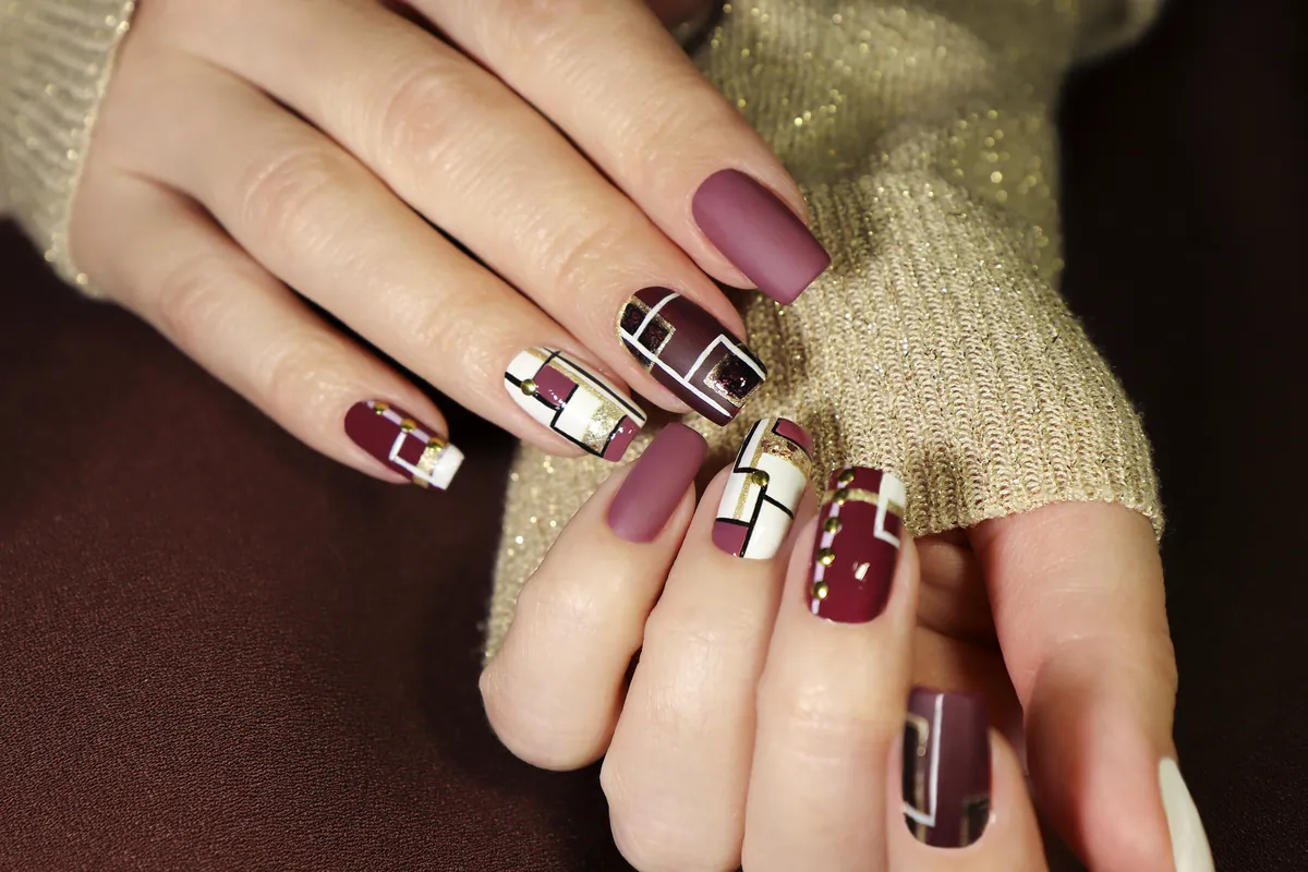 Nails in burgundy, mauve and white with geometric nail art against a beige knit