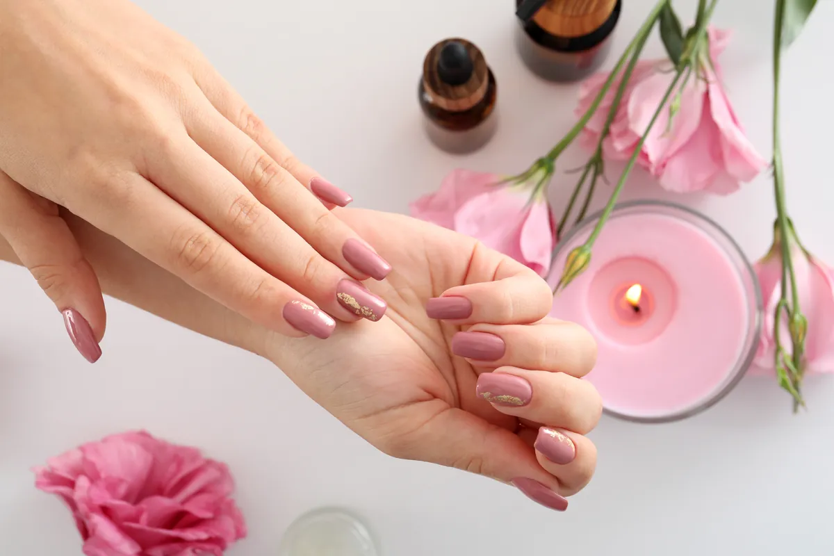 Hands with a soft pink manicure beside a candle, roses and small bottles of care oil