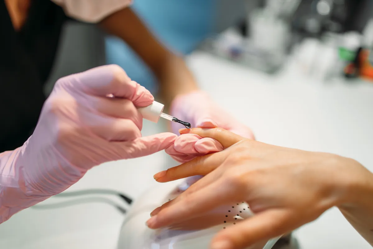 A nail technician in gloves applying polish to a client’s nails at a salon table