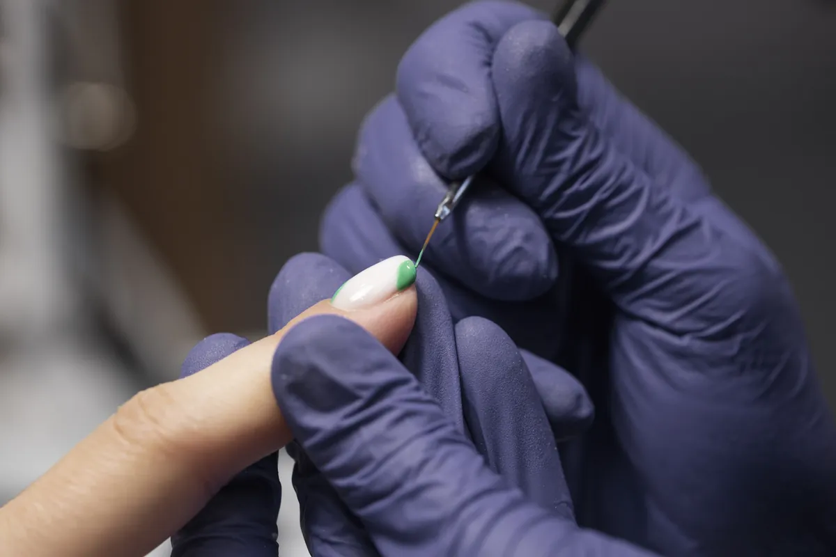 A nail artist in purple gloves painting a fine detail on a client’s nail with a thin brush
