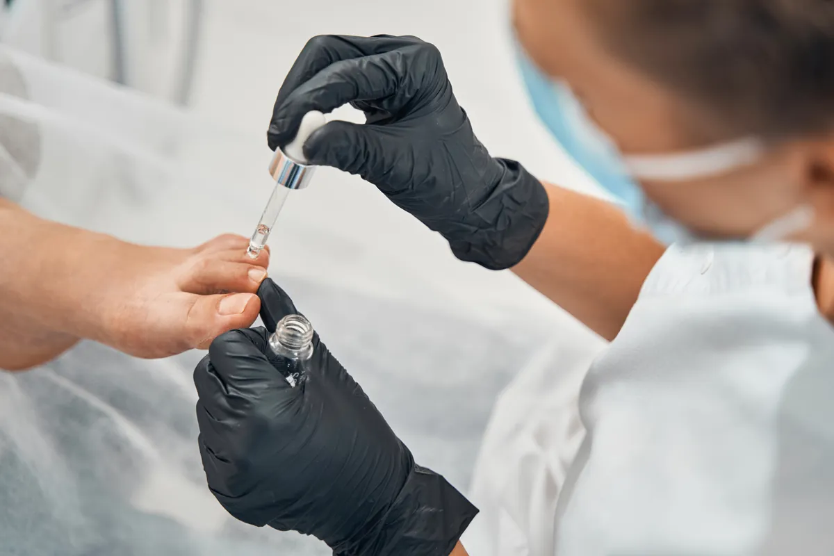 A nail technician in black gloves applying oil with a dropper to a client’s nails