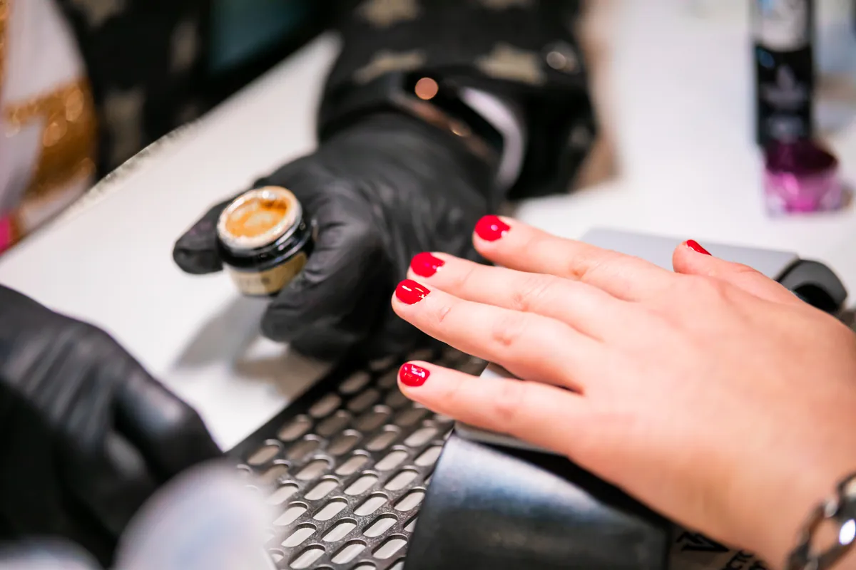 A technician in black gloves working on a client’s red manicure in a salon